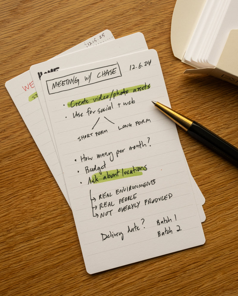 A notepad with handwritten meeting notes and a pen rests alongside a few Analog Lined Cards - Small. The notes mention creating video/photo assets, social/web use, quantity, budget, locations, real environments, and delivery dates. Some lines are highlighted yellow and green.