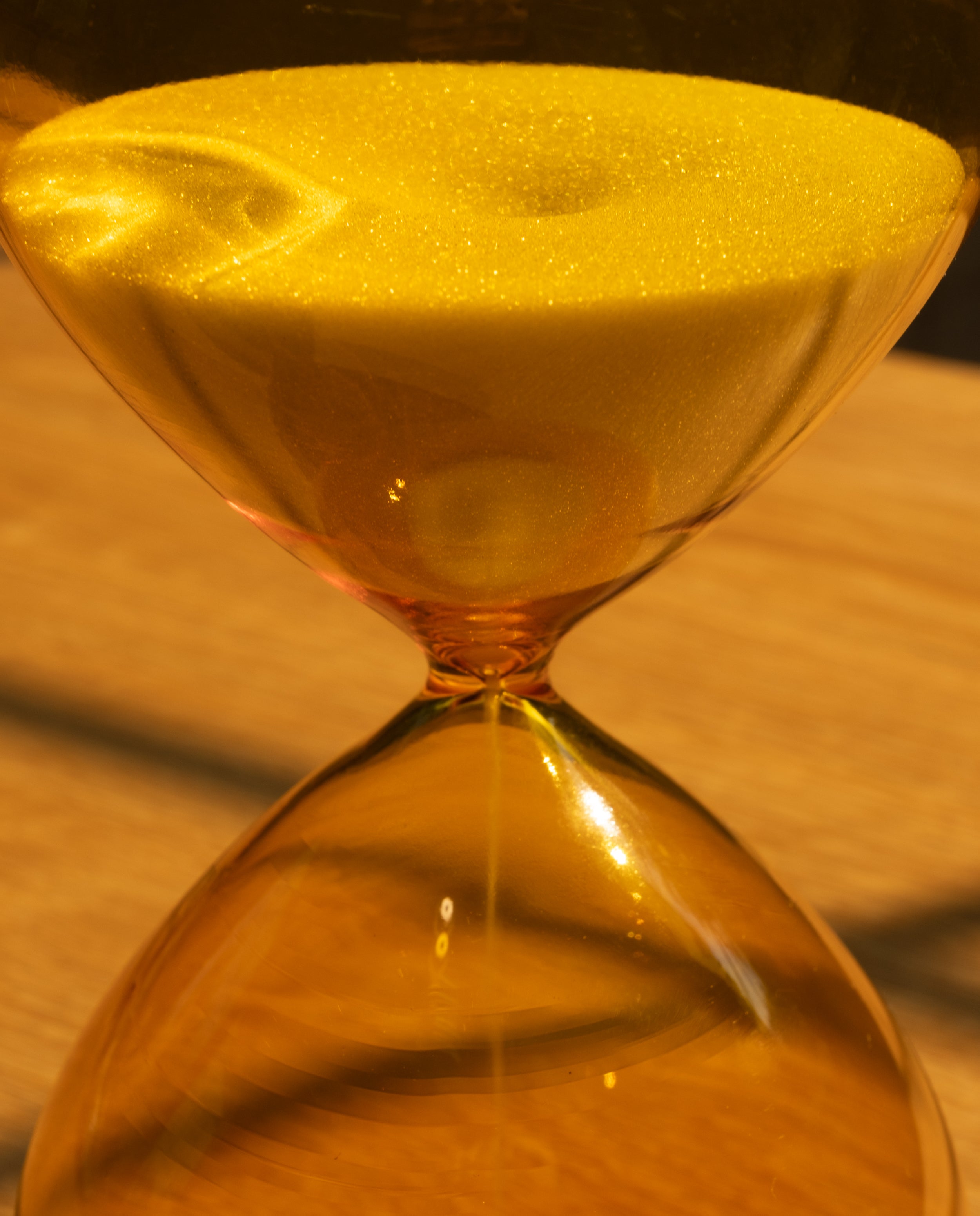 Close-up of the 25 Minute Hourglass (Amber), a hand-blown glass timer with yellow sand flowing between chambers, set on a wooden surface under warm lighting.