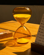 The 25 Minute Hourglass (Amber), a hand-blown glass timer with yellow sand, sits on a wooden desk in warm sunlight beside a pen and notepad.