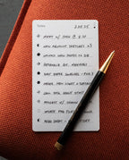 A premium Analog Trial Pack—including a handwritten to-do list on quality card stock and a black and gold pen—rests on an orange surface with tasks like meetings, research, photo uploads, and podcast recording displayed elegantly.