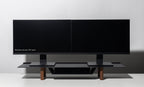 Two black 24-inch computer monitors are side by side on a Gather Large Monitor Stand (Black/Walnut) with wooden legs, set against a white background. The left monitor displays the text: “Monitors shown: 24” each.”.