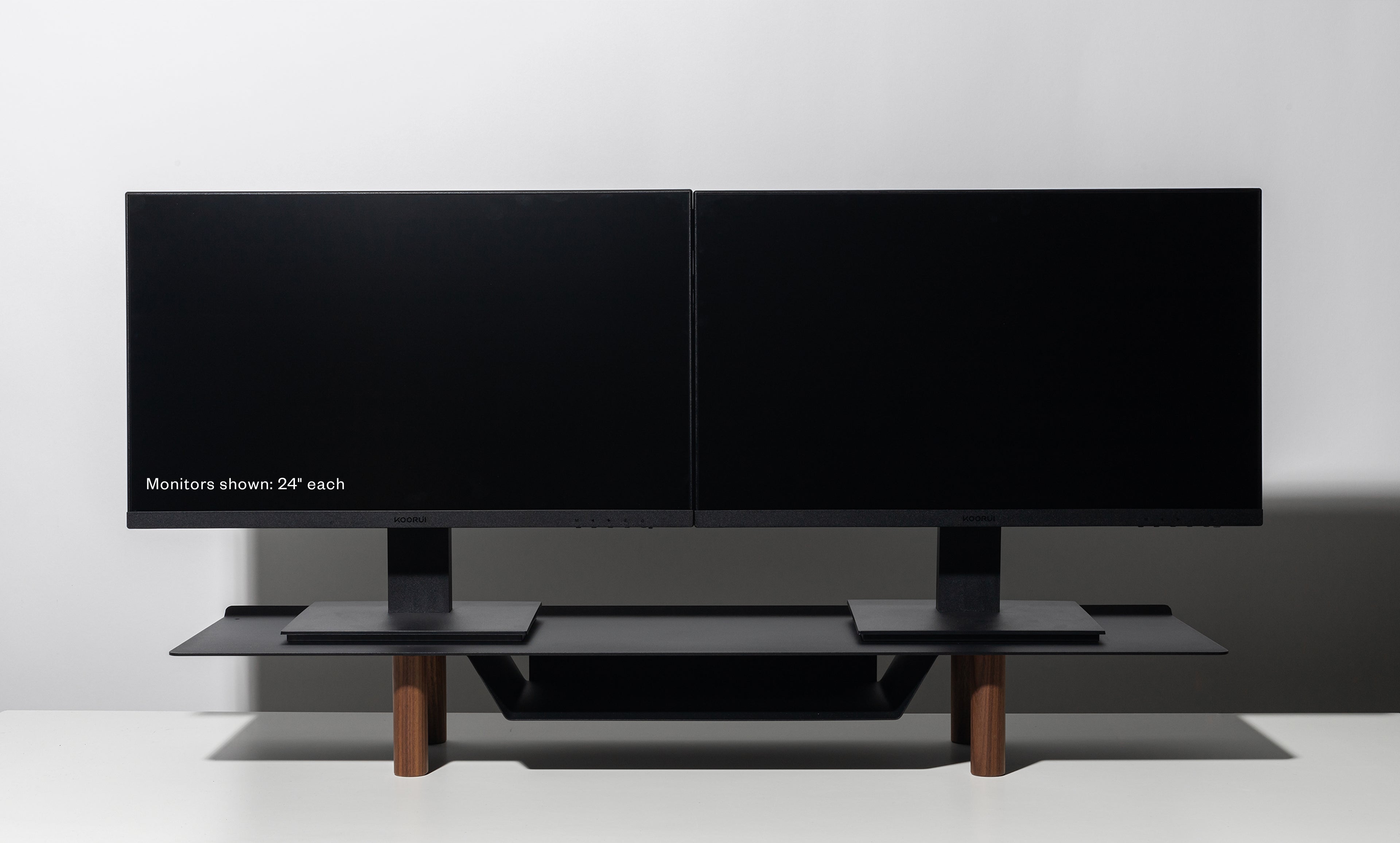 Two black 24-inch computer monitors are side by side on a Gather Large Monitor Stand (Black/Walnut) with wooden legs, set against a white background. The left monitor displays the text: “Monitors shown: 24” each.”.