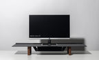 A 21-inch computer monitor sits on the Gather Large Monitor Stand (Black/Walnut) against a plain light gray background. The screen is off, drawing attention to the stand’s sleek, minimalist design and space-saving functionality.