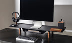 A minimalist black desk features the Gather Large Monitor Stand (Black/Walnut), with a monitor, keyboard, mouse, headphones on a holder, pen, and a compact desk organizer for stationery, all neatly arranged on a clean workspace.