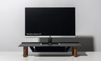 A 21-inch computer monitor rests on the Gather Small Monitor Stand (Black/Walnut) with wooden legs, set against a plain light gray background. The screen is off, displaying the text: “Monitor shown: 21”.”.