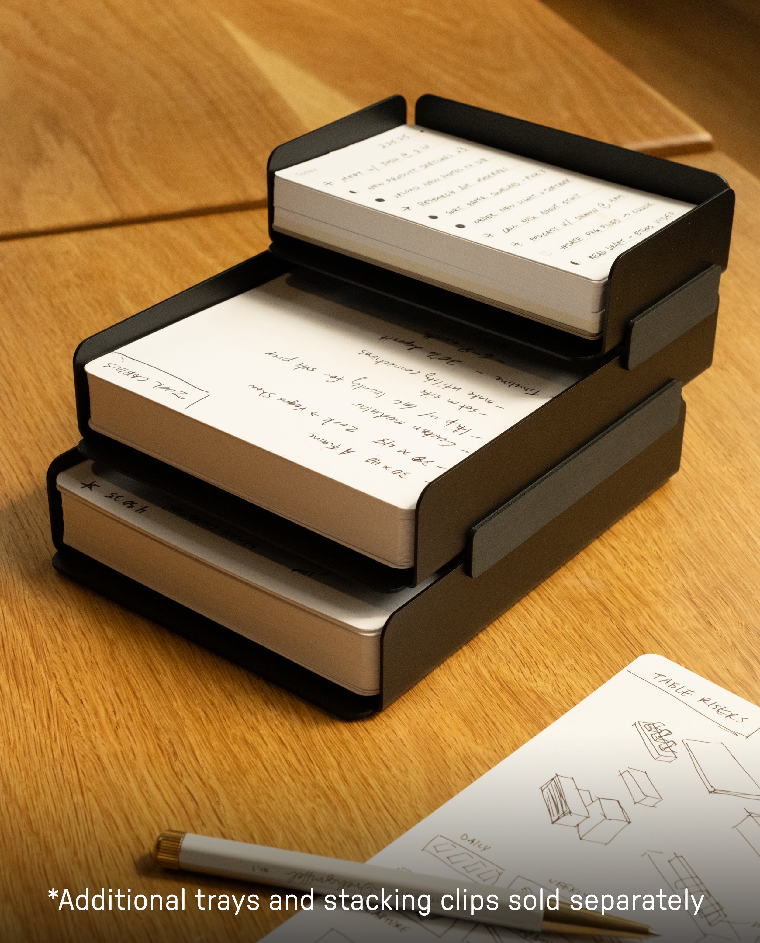 A black steel Capture Bundle (Small Tray + Large Tray) holds index cards with handwritten notes on a wooden desk. More Capture Cards and a pen are nearby. Text at the bottom: Additional trays and stacking clips sold separately.