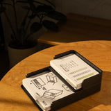 The Analog Capture Kit - Large (Black Aluminum) features illustrated dot grid cards in a sleek black holder on a wooden table, paired with a pen and open notebook. A potted plant adds to the creative workspace.