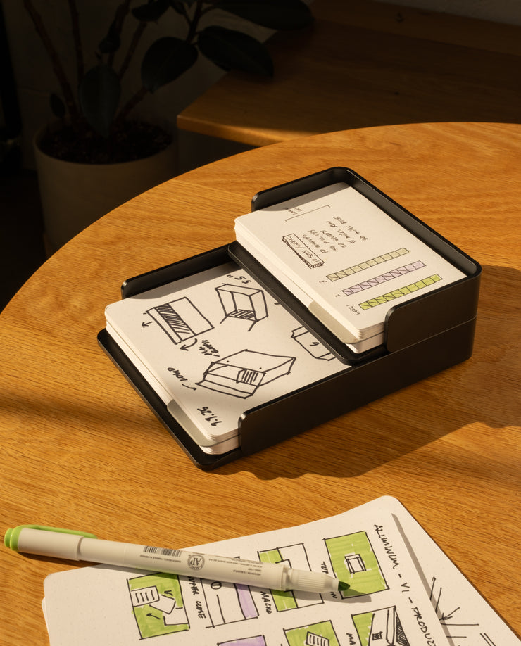 The Analog Capture Kit - Large (Black Aluminum) features illustrated dot grid cards in a sleek black holder on a wooden table, paired with a pen and open notebook. A potted plant adds to the creative workspace.