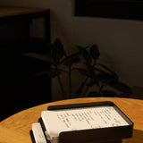 The Analog Capture Kit - 2 Large (Black Aluminum) sits on a round wooden table with dot grid cards and a black pen, bathed in sunlight with warm shadows and a blurred plant in the background.