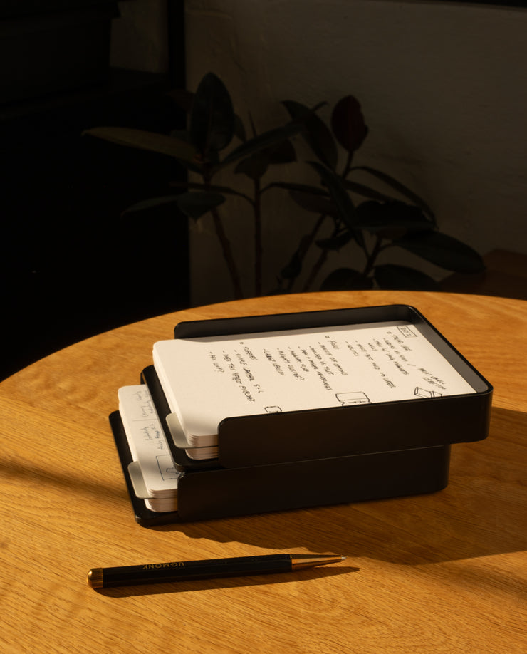 The Analog Capture Kit - 2 Large (Black Aluminum) sits on a round wooden table with dot grid cards and a black pen, bathed in sunlight with warm shadows and a blurred plant in the background.