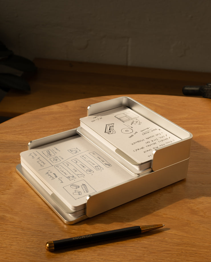 The Analog Capture Kit - Small (Silver Aluminum) with dot grid cards sits on a wooden table. A black and gold pen lies in front, while warm sunlight highlights the inviting setup, perfect for capturing hand-drawn diagrams and notes.