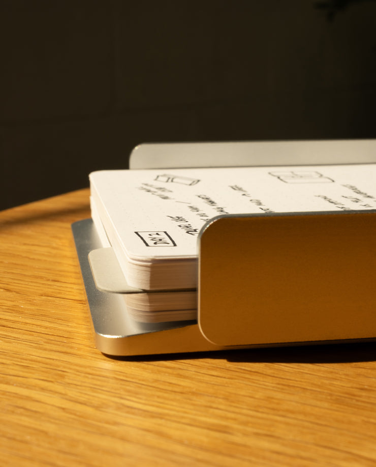 A close-up of the Analog Capture Kit - Large (Silver Aluminum) shows white dot grid cards with sketches and writing, stacked in a sleek silver aluminum tray on a wooden surface under warm light.