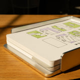 The Analog Capture Kit - Large (Silver Aluminum) rests on a wooden desk, its dot grid cards with green diagrams neatly stacked in the aluminum tray, sunlight casting shadows. A pen appears in the foreground, ready for organized note capture.