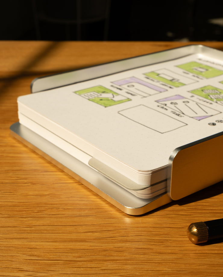 The Analog Capture Kit - Large (Silver Aluminum) rests on a wooden desk, its dot grid cards with green diagrams neatly stacked in the aluminum tray, sunlight casting shadows. A pen appears in the foreground, ready for organized note capture.