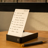 The Analog Daily Focus Kit (Black Aluminum) sits on a wooden desk, displaying a handwritten to-do list in its sleek black holder, accompanied by task cards and a black and gold pen. A window with black framing suggests an organized workspace.