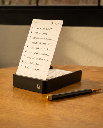 The Analog Daily Focus Kit (Black Aluminum) sits on a wooden desk, displaying a handwritten to-do list in its sleek black holder, accompanied by task cards and a black and gold pen. A window with black framing suggests an organized workspace.
