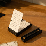A notepad with a handwritten to-do list stands upright in The Complete Analog Collection (Black Aluminum) organizer on a wooden desk, next to a matching black pen holder. Loose note sheets and warm sunlight complete the scene.