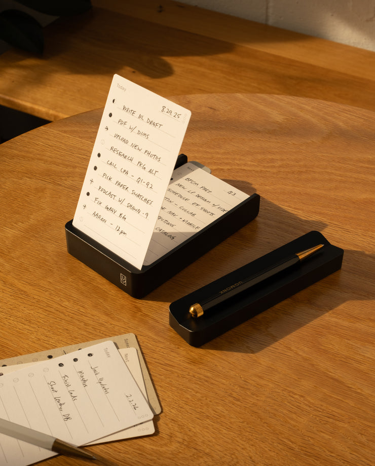 A notepad with a handwritten to-do list stands upright in The Complete Analog Collection (Black Aluminum) organizer on a wooden desk, next to a matching black pen holder. Loose note sheets and warm sunlight complete the scene.