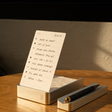 The Complete Analog Collection (Silver Aluminum) holds a handwritten to-do list upright on a wooden table, with a black and gold pen resting in its matching tray, all beautifully illuminated by warm sunlight.