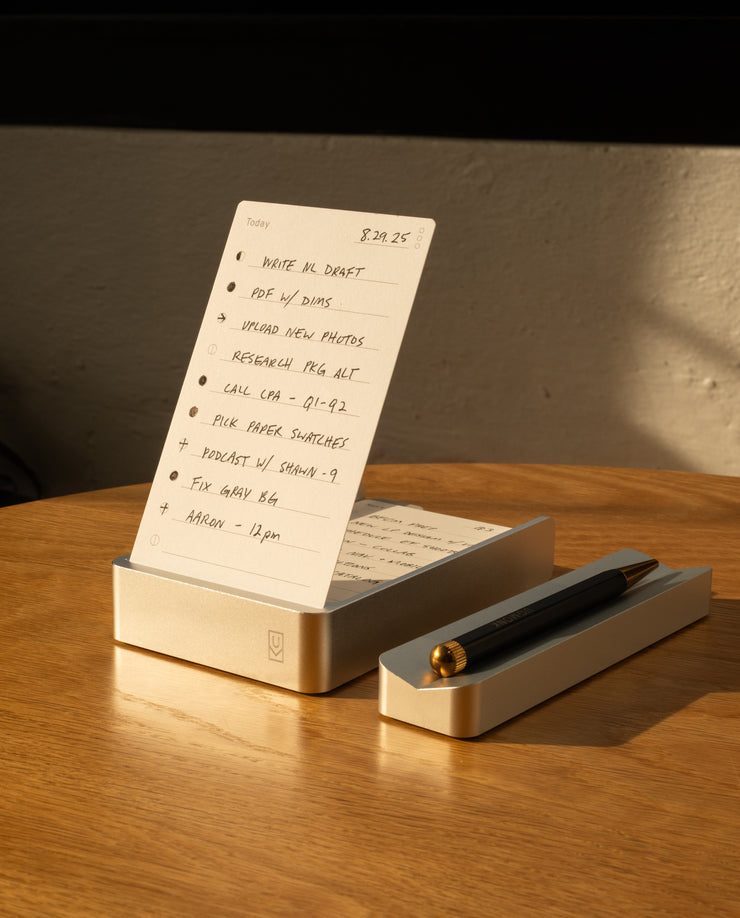 The Complete Analog Collection (Silver Aluminum) holds a handwritten to-do list upright on a wooden table, with a black and gold pen resting in its matching tray, all beautifully illuminated by warm sunlight.