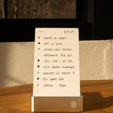 A handwritten to-do list on the Analog Daily Focus Kit + 6 Refills (Silver Aluminum) rests on a wooden table in sunlight, listing tasks like drafting, calling CPA, and a 9am podcast. A pen and notebook nearby highlight this analog productivity system.