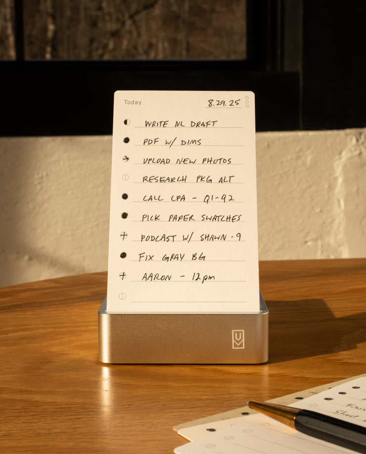 A handwritten to-do list on the Analog Daily Focus Kit + 6 Refills (Silver Aluminum) rests on a wooden table in sunlight, listing tasks like drafting, calling CPA, and a 9am podcast. A pen and notebook nearby highlight this analog productivity system.