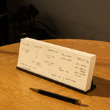 An Analog Weekly Planning Kit (Black Aluminum) sits on a wooden table beside a black and gold pen and an Aluminum Card Stand, sunlight casting shadows as the planner displays daily tasks and appointments.