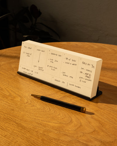 An Analog Weekly Planning Kit (Black Aluminum) sits on a wooden table beside a black and gold pen and an Aluminum Card Stand, sunlight casting shadows as the planner displays daily tasks and appointments.
