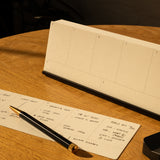 The Analog Weekly Planning Kit (Black Aluminum) includes Weekly Cards and an aluminum card stand for organization, shown here with a blank desk calendar and a black and gold pen on a handwritten planner sheet atop a wooden table.