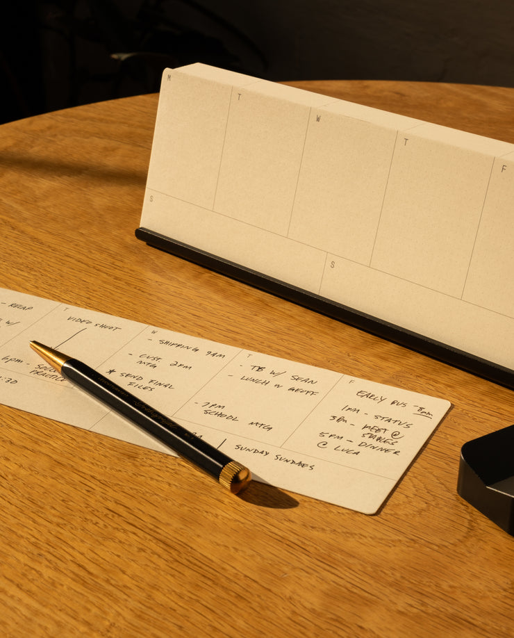 The Analog Weekly Planning Kit (Black Aluminum) includes Weekly Cards and an aluminum card stand for organization, shown here with a blank desk calendar and a black and gold pen on a handwritten planner sheet atop a wooden table.