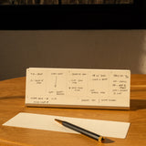 An Analog Weekly Planning Kit (Silver Aluminum) with handwritten notes sits on a round wooden table beside a notepad, black and gold pen, and aluminum card stand, as sunlight streams in from a nearby window, casting warm light and shadows.