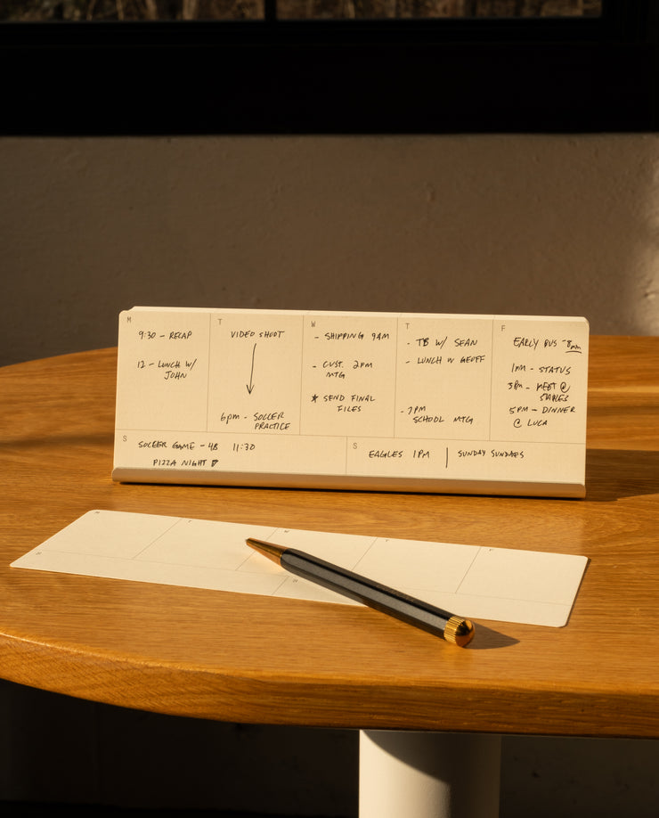 An Analog Weekly Planning Kit (Silver Aluminum) with handwritten notes sits on a round wooden table beside a notepad, black and gold pen, and aluminum card stand, as sunlight streams in from a nearby window, casting warm light and shadows.