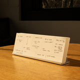 An Analog Weekly Planning Kit (Silver Aluminum) sits open on a wooden table in warm sunlight, propped up by its aluminum card stand and displaying schedules in black ink. A dark window and wall appear in the background.