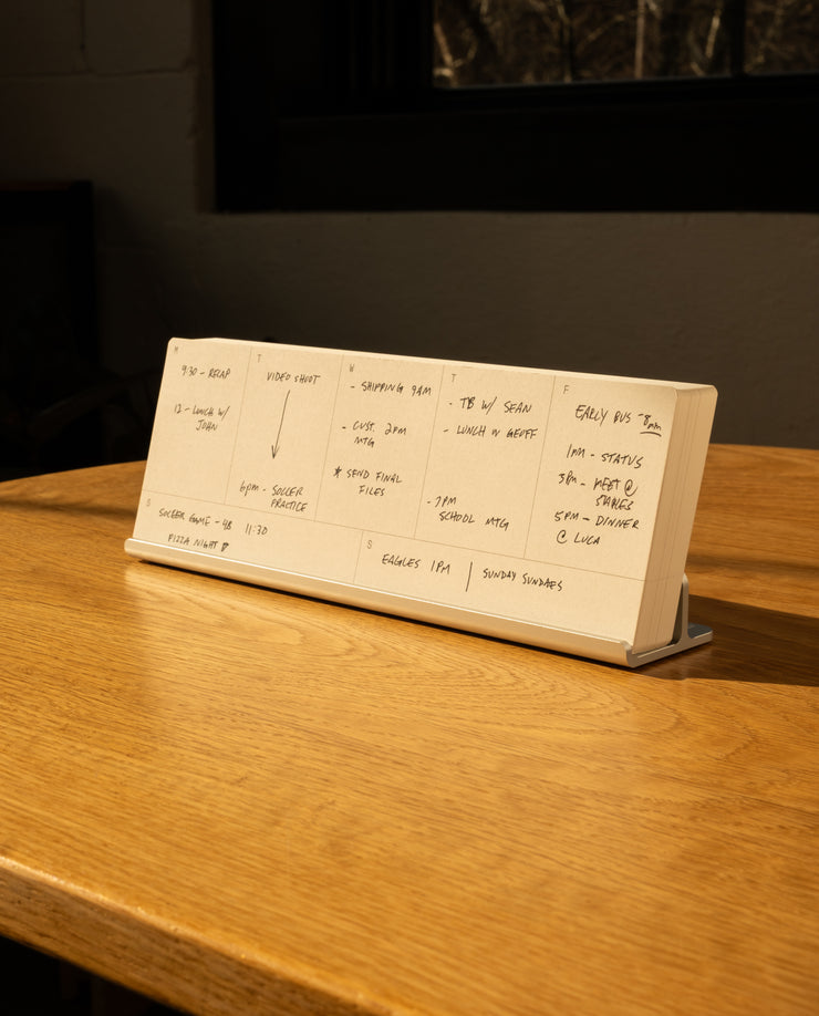 An Analog Weekly Planning Kit (Silver Aluminum) sits open on a wooden table in warm sunlight, propped up by its aluminum card stand and displaying schedules in black ink. A dark window and wall appear in the background.