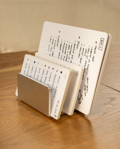 The Complete Analog Collection (Silver Aluminum) minimalist metal desktop organizer neatly holds multiple open notebooks of various sizes, filled with handwritten notes and sketches, on a wooden surface.