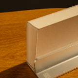 A thick paperback book stands upright in the Analog Weekly Planning Kit (Silver Aluminum) on a wooden surface, casting a soft shadow—ideal for keeping your week’s plans and cards organized and easily visible.