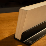 An Analog Weekly Planning Kit (Black Aluminum) stands vertically in its holder on a wooden surface, while warm sunlight casts gentle shadows.