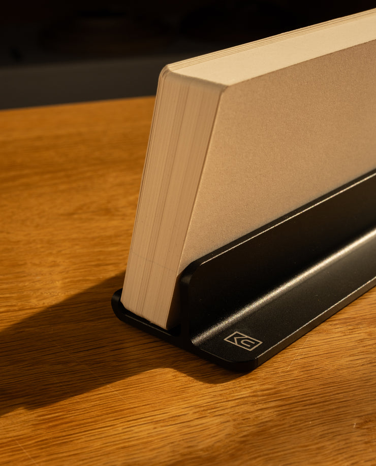 An Analog Weekly Planning Kit (Black Aluminum) stands vertically in its holder on a wooden surface, while warm sunlight casts gentle shadows.