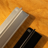 Two metal objects, one silver and one black with geometric logos on their ends, rest on wood. Warm light casts shadows, evoking the sleek design of the Analog Weekly Planning Kit (Silver Aluminum).