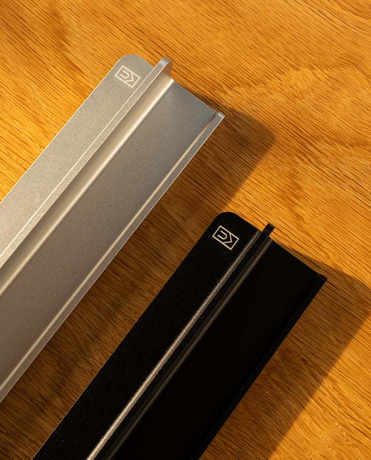 Two metal objects, one silver and one black with geometric logos on their ends, rest on wood. Warm light casts shadows, evoking the sleek design of the Analog Weekly Planning Kit (Silver Aluminum).