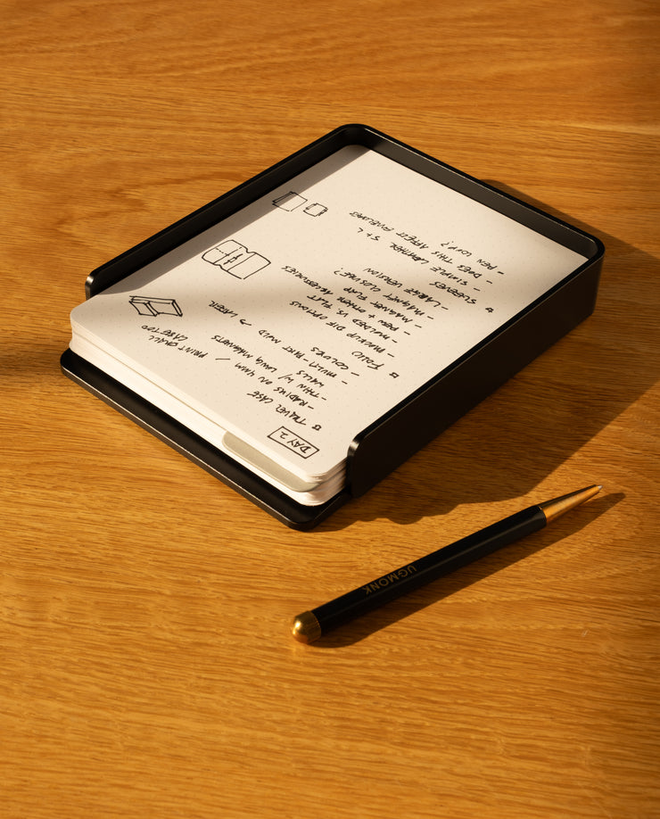 The Analog Capture Kit - Large (Black Aluminum) features a black aluminum card tray holding dot grid cards with handwritten notes and sketches. A black pen with a gold tip sits on a light wooden surface next to the kit.