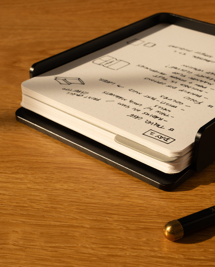The Analog Capture Kit - Large (Black Aluminum) includes large dot grid cards in a sleek black holder, with a black pen featuring a gold tip, all resting on a wooden surface—an ideal setup for your notes.
