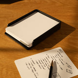 The Analog Capture Kit - Small & Large (Black Aluminum) with blank dot grid cards in a black holder rests on a wooden surface beside a handwritten note and pen, while sunlight casts shadows across the scene.