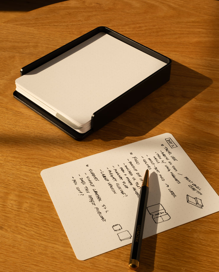 The Analog Capture Kit - Small & Large (Black Aluminum) with blank dot grid cards in a black holder rests on a wooden surface beside a handwritten note and pen, while sunlight casts shadows across the scene.