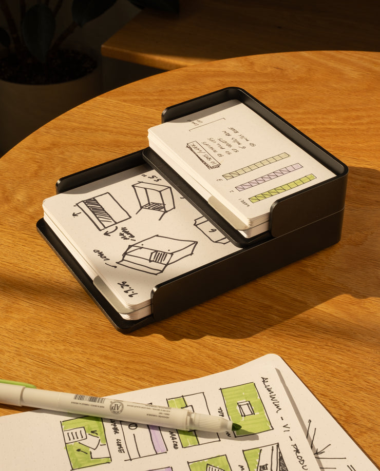 The Analog Capture Kit - Small & Large (Black Aluminum) rests on a wooden table beside an open notebook with colorful sketches and a green marker, as sunlight casts warm shadows over this creative setup.