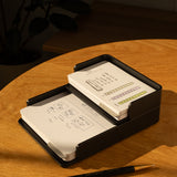 The Complete Analog Collection (Black Aluminum) two-tier card organizer sits on a wooden table holding index cards with diagrams and handwritten notes. Nearby is a black and gold pen, with a blurred plant in the background.