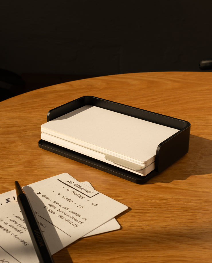 The Complete Analog Collection (Black Aluminum) sits on a wooden table with blank index cards in a sleek black organizer, beside a handwritten note and black pen as warm sunlight casts soft shadows over the scene.