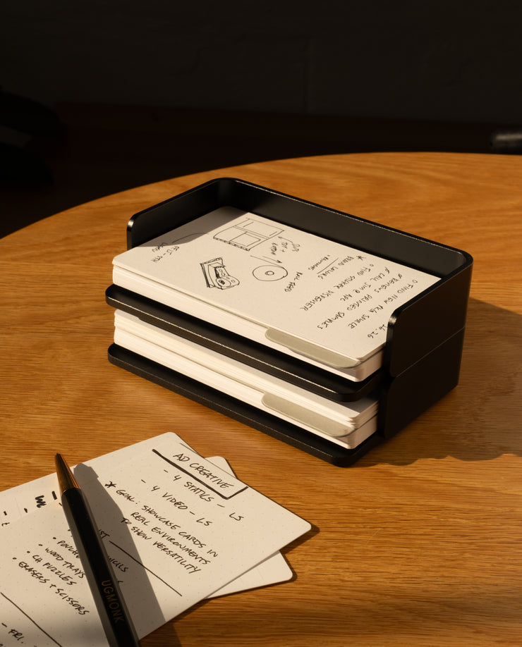 An Analog Capture Kit - Small (Black Aluminum) sits on a wooden table, holding Dot Grid Cards. The top card features sketches and notes; nearby, a pen and two more notecards from the kit with handwritten notes are partly visible.