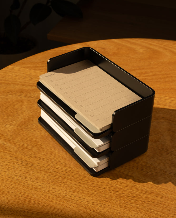 A black Analog Capture Kit - Small (Black Aluminum) holds stacks of brown and white Dot Grid Cards on a wooden table in warm sunlight.