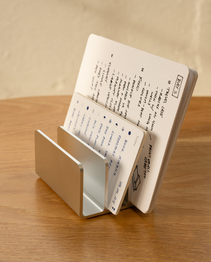 The Analog Card Organizer (Silver Aluminum) holds an open notebook with handwritten notes and sketches, displayed on a wooden surface against a beige background.